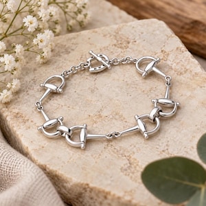 May include: A silver-coloured chain bracelet featuring repeating equestrian-inspired bit links. The bracelet is constructed with polished metal components connected by small circular rings, ending in a toggle clasp mechanism with a horseshoe-shaped bar.