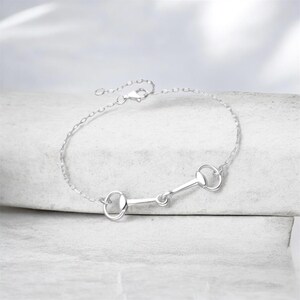 May include: A silver-toned bracelet featuring a central horse bit motif connected to a delicate chain. The piece includes a small lobster claw clasp and a short extender chain, showcasing a polished metallic finish throughout the entire design.