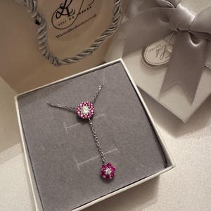 May include: A silver necklace with a floral pendant featuring pink and white gemstones, presented in a white gift box with a grey interior. A matching gift bag and box with a silver bow are also visible.