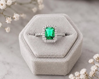 Puede incluir: Un anillo de tono plateado con una piedra central verde rectangular de corte esmeralda rodeada por un halo de pequeñas piedras blancas. La banda también está engastada con pequeñas piedras blancas en los laterales. El anillo se presenta en un estuche hexagonal de terciopelo gris.