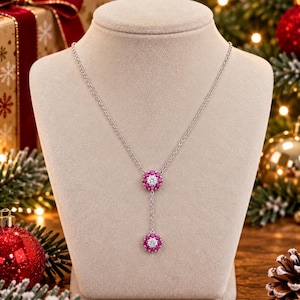 Puede incluir: Un collar de tono plateado con dos colgantes circulares, cada uno diseñado con una piedra central transparente rodeada por un anillo de pequeñas piedras de color rosa vibrante. Los colgantes están unidos por una fina cadena plateada en forma de Y.
