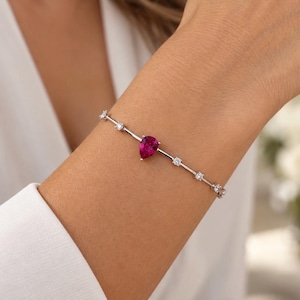 May include: A silver-toned bracelet featuring a central pear-shaped deep pink gemstone flanked by several small, round, clear sparkling stones set along a thin, delicate metal chain that wraps around the wrist with a refined and polished appearance.