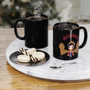 May include: Two black coffee mugs with steam rising, one plain and one with the word "APSARA" and a cartoon graphic. A black plate holds macarons and chocolate sticks. A silver spoon rests on the marble tray.