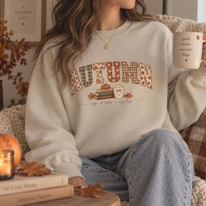 Puede incluir: Una sudadera beige de cuello redondo con la palabra AUTUMN en una fuente de patrones variados con gráficos de temática otoñal que incluyen una calabaza, una pila de libros y una taza, con el texto coffee, books, cozy days impreso debajo.