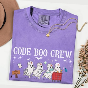 Puede incluir: Una camiseta morada con un estampado gráfico de cuatro figuras fantasmales vestidas como profesionales médicos. El texto dice CODE BOO CREW y BOO WE SAVE YOU. La camiseta tiene una etiqueta Comfort Colors y un collar de estrella dorada.