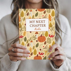 Könnte beinhalten: Ein Notizbuch mit festem Einband auf cremefarbenem Hintergrund mit einem Muster aus orangefarbenen Kürbissen, roten Beeren, braunen Eicheln sowie verschiedenen grünen und gelben Blättern. Ein weißes Etikett zeigt den Text MY NEXT CHAPTER und Amelia.