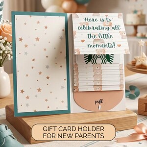 May include: A gift card holder for new parents. The card holder is open, revealing a Starbucks gift card and a pull tab. The card holder has a teal border and a star pattern. The text "Here is to celebrating all the little moments!" is visible.