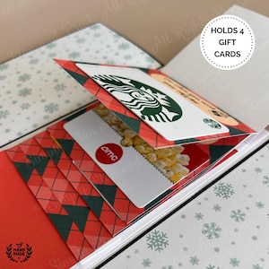 May include: A handmade gift card holder with a red and green plaid design. The holder is open, revealing gift cards from Starbucks and AMC. The holder is designed to hold four gift cards. The background has a snowflake pattern.