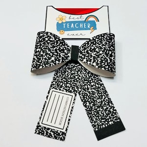 May include: A decorative bow featuring a composition book design with black and white speckled paper. The bow has a black center and two long streamers. The top portion reads "Best Teacher Ever" with a rainbow and lightning bolt.