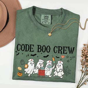 May include: A sage green t-shirt featuring the text CODE BOO CREW in black letters. The graphic displays four white ghosts dressed as medical staff with a medical cart, a syringe, a clipboard, a pumpkin, bats, and a spider web in black and orange.