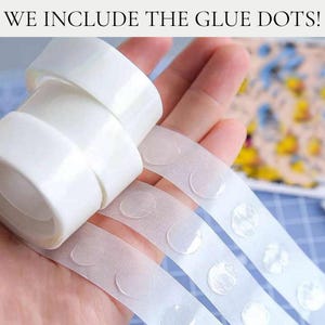 May include: A roll of clear adhesive glue dots on a white backing strip with the text WE INCLUDE THE GLUE DOTS! visible at the top. The transparent circular adhesive dots are arranged in a single row on the peelable white paper tape.