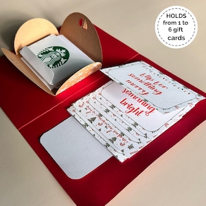 May include: A red gift card holder with a brown card holder inside, holding a Starbucks gift card. The holder also contains several gift cards with a festive design and the text "flip for something merry, something bright". The holder can hold from 1 to 6 gift cards.