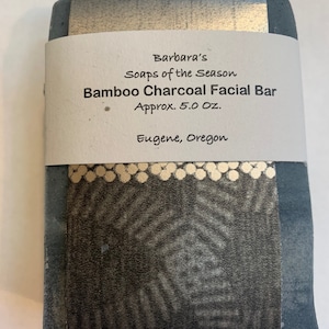 Op de afbeelding: Een stuk houtskoolzeep met een wit etiket waarop staat "Barbara's Soaps of the Season Bamboo Charcoal Facial Bar Approx. 5.0 Oz. Eugene, Oregon".