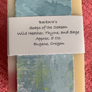 May include: A bar of soap wrapped in paper with a blue and green design. The paper has text that reads "Barbara's Soaps of the Season Wild Heather, Thyme, and Sage Approx. 5 Oz. Eugene, Oregon"