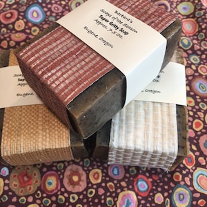 以下が含まれることがあります： 自然繊維の布で包まれた茶色の石鹸が3つ。石鹸には「Soaps of the Season Super Gritty Soap Approx. 7.5 Oz. Barbara's Eugene, Oregon」とラベルが貼られています