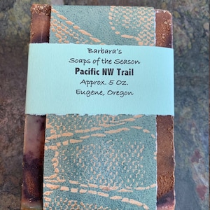 May include: A bar of soap with a brown and white swirl pattern. The soap is wrapped in a light blue paper with a gold design. The paper has the text "Barbara's Soaps of the Season Pacific NW Trail Approx. 5 Oz. Eugene, Oregon" printed on it.
