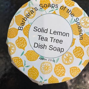 May include: Round dish soap container with a white and yellow lemon pattern. The label reads "Solid Lemon Tea Tree Dish Soap" and "Barbara's Soaps of the Season." The container is approximately 2.5 oz.