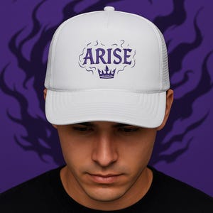 May include: White trucker hat with a curved brim and mesh back. The front of the hat features the word "ARISE" in purple script above a purple crown design. The hat is worn by a person.