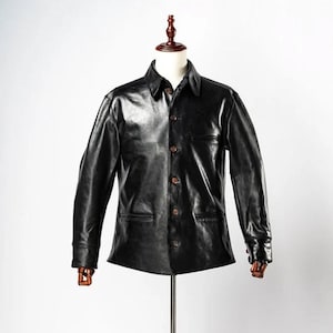 Puede incluir: Chaqueta de cuero negro con cuello y cierre de botones. La chaqueta tiene dos bolsillos delanteros y mangas largas. La chaqueta se exhibe en un maniquí con un soporte de madera.