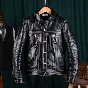 May include: A black leather jacket with a classic design, featuring a collared neckline, button-down front, and two chest pockets. The jacket has gold stitching and is hanging on a wooden hanger.