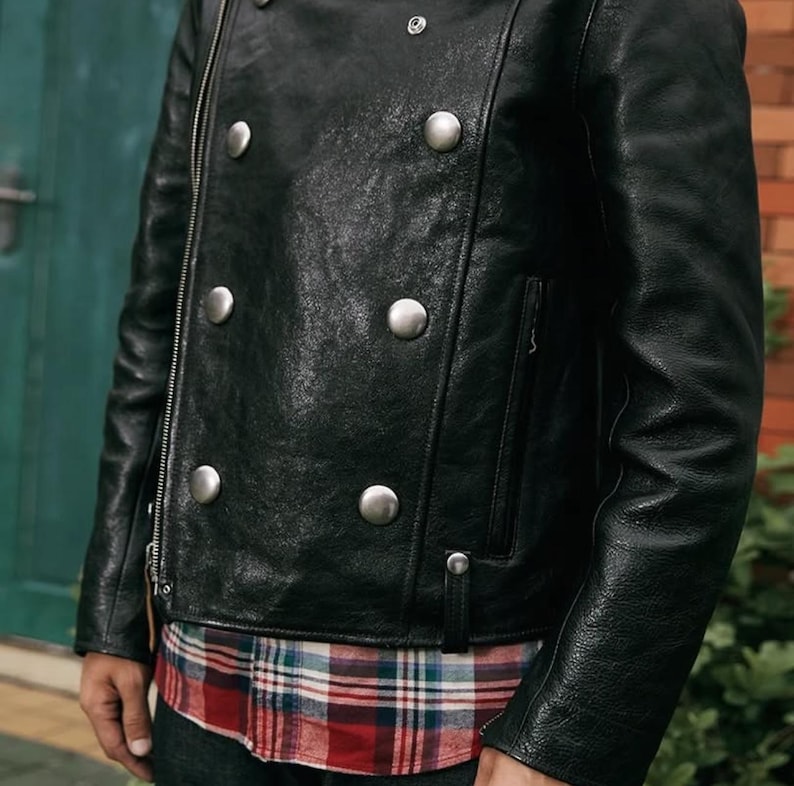 Pode incluir: Casaco de couro preto com grandes bot&otilde;es prateados e fecho de correr. O casaco tem um estilo motociclista cl&aacute;ssico com um design de abotoamento duplo. Uma camisa xadrez vermelha, branca e azul &eacute; vis&iacute;vel por baixo.