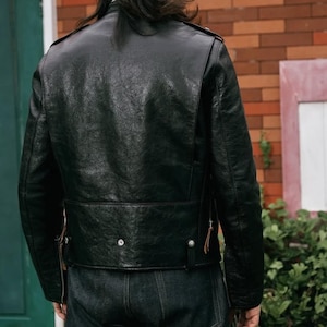 Pode incluir: Casaco de couro preto com acabamento texturizado. O casaco apresenta um estilo motociclista cl&aacute;ssico com gola entalhada, ombreiras e cinto na cintura. A parte de tr&aacute;s do casaco &eacute; vis&iacute;vel, mostrando os detalhes do design.
