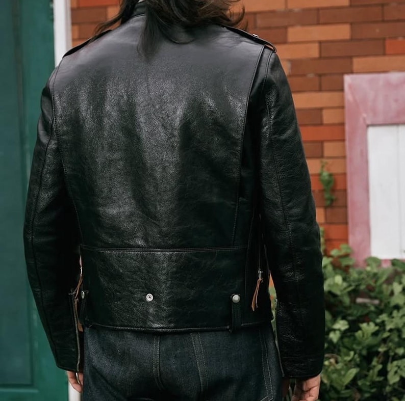 Pode incluir: Casaco de couro preto com acabamento texturizado. O casaco apresenta um estilo motociclista cl&aacute;ssico com gola entalhada, ombreiras e cinto na cintura. A parte de tr&aacute;s do casaco &eacute; vis&iacute;vel, mostrando os detalhes do design.