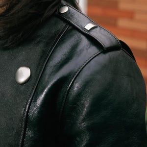 Pode incluir: Close-up de uma jaqueta de couro preta. A jaqueta apresenta bot&otilde;es e detalhes nos ombros em metal prateado. O couro tem um acabamento brilhante e detalhes de costura vis&iacute;veis. Um estilo de jaqueta cl&aacute;ssico.