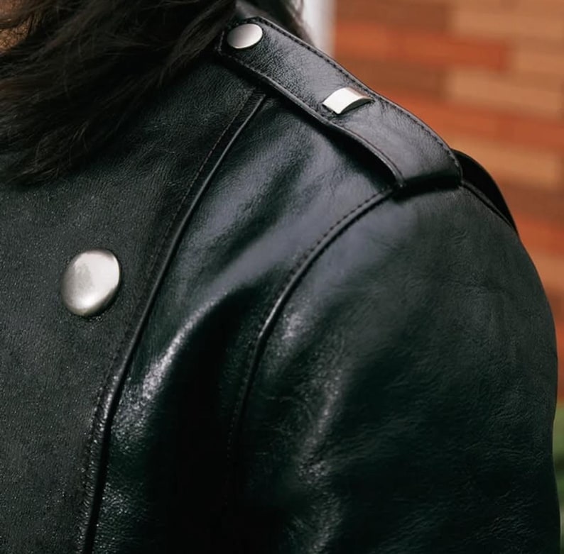 Pode incluir: Close-up de uma jaqueta de couro preta. A jaqueta apresenta bot&otilde;es e detalhes nos ombros em metal prateado. O couro tem um acabamento brilhante e detalhes de costura vis&iacute;veis. Um estilo de jaqueta cl&aacute;ssico.