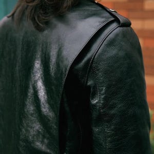 Pode incluir: Um close-up de uma jaqueta de couro verde escuro. A jaqueta tem uma superf&iacute;cie texturizada e um detalhe no ombro com uma pequena al&ccedil;a e um bot&atilde;o. A jaqueta parece estar bem usada.