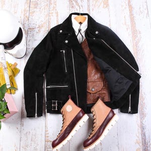 May include: A black suede motorcycle jacket with silver zippers and a brown leather vest. A pair of two-tone leather boots with white soles and a white motorcycle helmet are also visible. The items are arranged on a light wood surface.