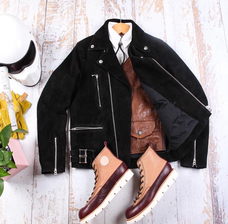 May include: A black suede motorcycle jacket with silver zippers and a brown leather vest. A pair of two-tone leather boots with white soles and a white motorcycle helmet are also visible. The items are arranged on a light wood surface.