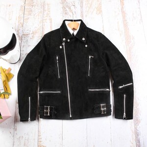 May include: Black suede motorcycle jacket with silver zippers and buckles. The jacket has a classic biker style with multiple pockets and a notched collar. It is hanging on a wooden hanger against a light wood background.