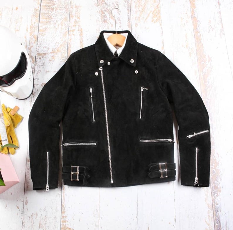 May include: Black suede motorcycle jacket with silver zippers and buckles. The jacket has a classic biker style with multiple pockets and a notched collar. It is hanging on a wooden hanger against a light wood background.