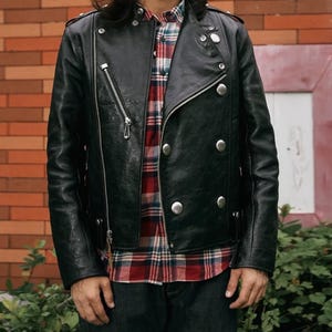 Pode incluir: Jaqueta de couro preta com z&iacute;per diagonal e bot&otilde;es prateados. A jaqueta tem um estilo motociclista cl&aacute;ssico com gola entalhada e v&aacute;rios bolsos. O modelo est&aacute; vestindo uma camisa xadrez vermelha e preta por baixo.