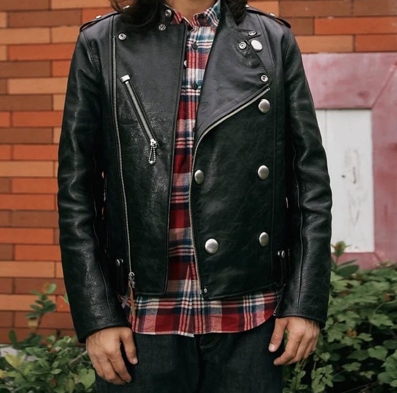 Pode incluir: Jaqueta de couro preta com z&iacute;per diagonal e bot&otilde;es prateados. A jaqueta tem um estilo motociclista cl&aacute;ssico com gola entalhada e v&aacute;rios bolsos. O modelo est&aacute; vestindo uma camisa xadrez vermelha e preta por baixo.