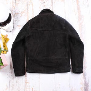 May include: A black suede jacket hangs on a metal hanger. The jacket has a classic design with a collar, a small pocket detail, and side adjustment straps. A white motorcycle helmet is visible in the background.