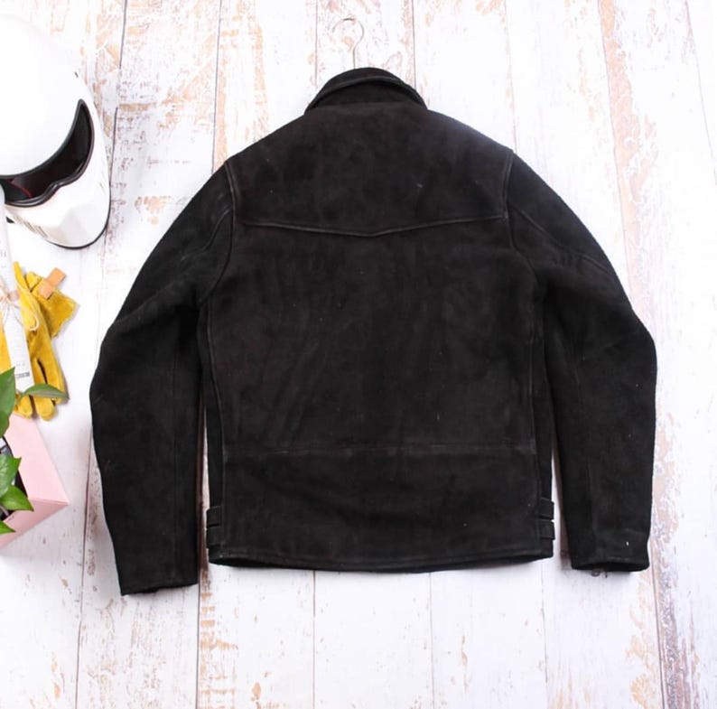 May include: A black suede jacket hangs on a metal hanger. The jacket has a classic design with a collar, a small pocket detail, and side adjustment straps. A white motorcycle helmet is visible in the background.