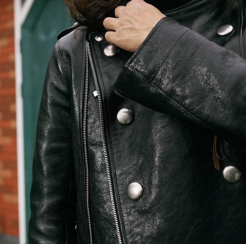 Pode incluir: Casaco de couro preto com z&iacute;per prateado e bot&otilde;es grandes prateados. O casaco tem um estilo motociclista cl&aacute;ssico com um acabamento texturizado. A imagem mostra um close-up dos detalhes do casaco, incluindo a gola e as mangas.
