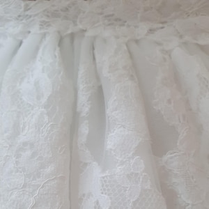 May include: Close-up of a white lace skirt. The skirt features intricate floral lace patterns over a sheer fabric. The waistband is also detailed with lace, creating a delicate and elegant look. Ideal for bridal or formal wear.