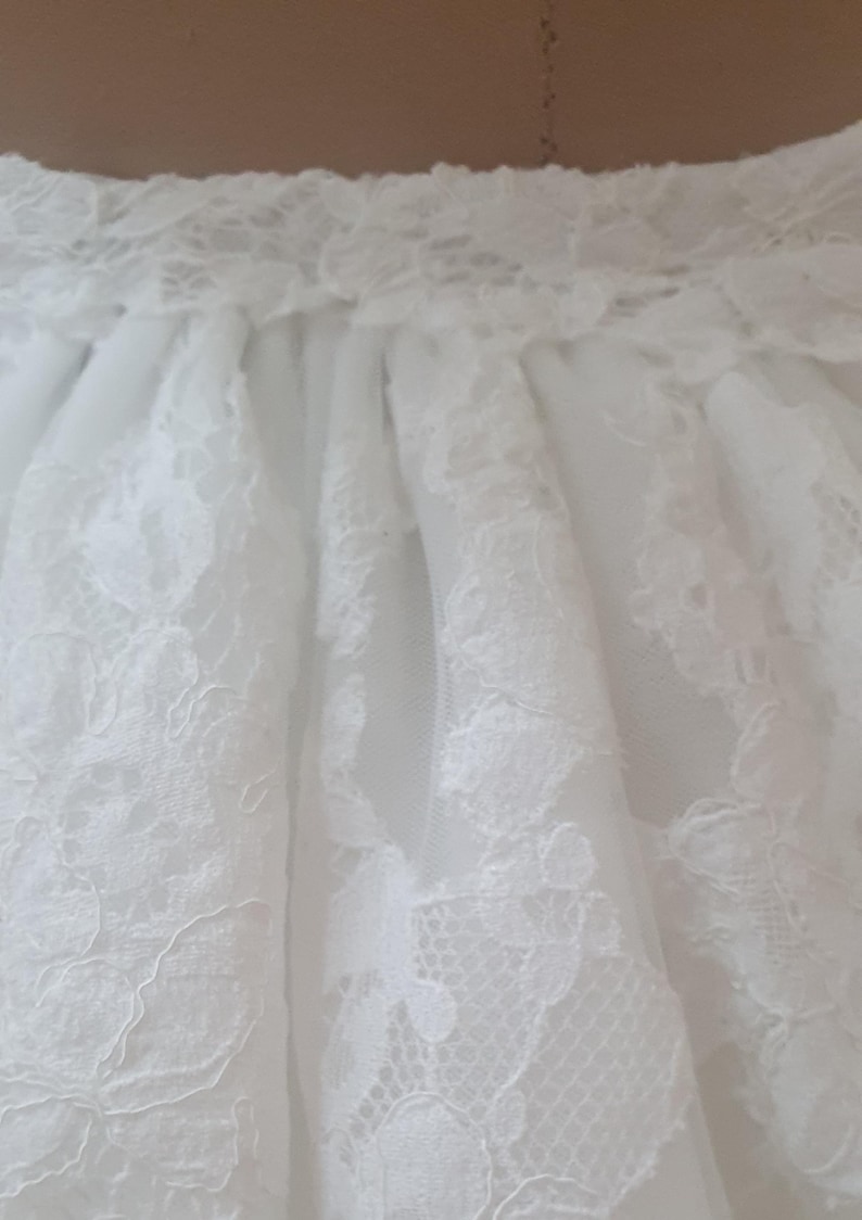 May include: Close-up of a white lace skirt. The skirt features intricate floral lace patterns over a sheer fabric. The waistband is also detailed with lace, creating a delicate and elegant look. Ideal for bridal or formal wear.
