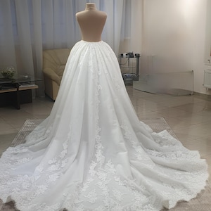 May include: A white wedding dress on a mannequin. The dress features a full skirt with intricate lace detailing along the hem and train. The fabric appears to be a soft, flowing material, creating a romantic and elegant look.