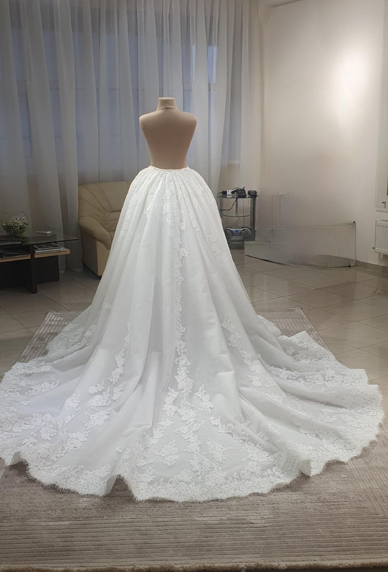 May include: A white wedding dress on a mannequin. The dress features a full skirt with intricate lace detailing along the hem and train. The fabric appears to be a soft, flowing material, creating a romantic and elegant look.