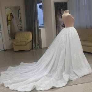 May include: A white wedding dress on a mannequin. The dress features a long train with intricate lace detailing along the hem. The mannequin is set in a room with a neutral color palette, with a mirror and a couch in the background.
