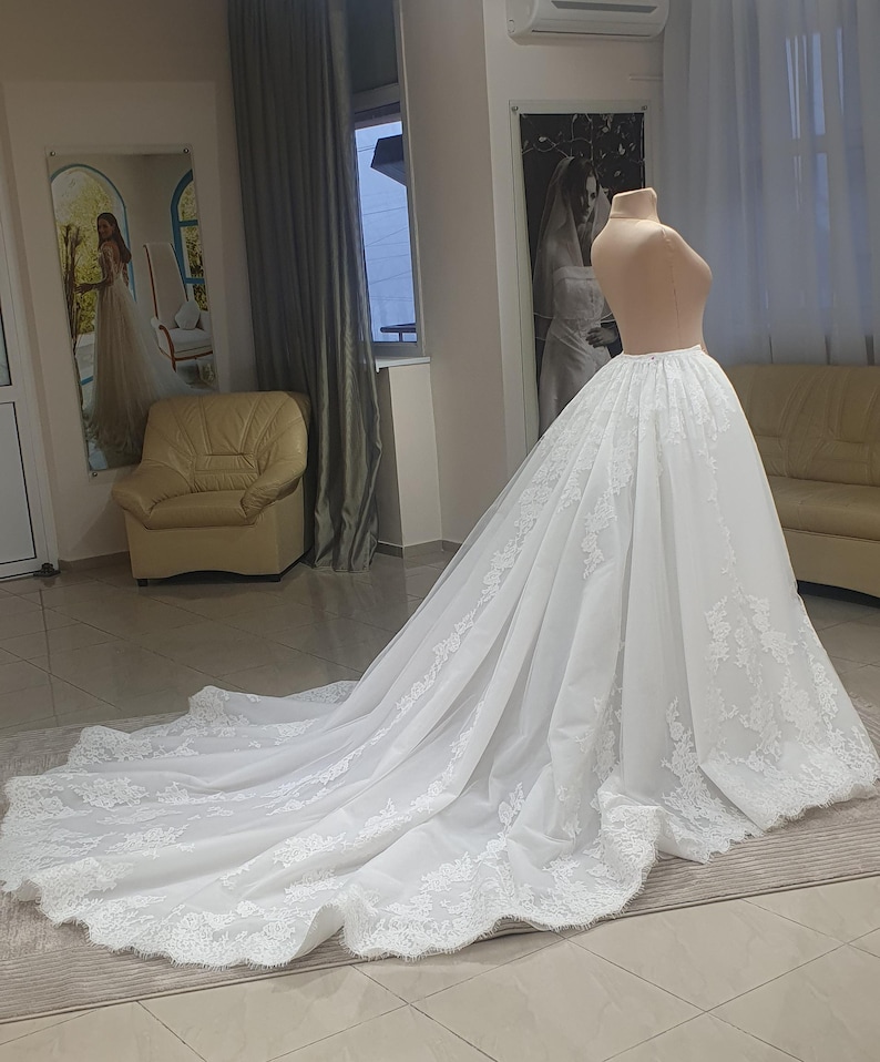 May include: A white wedding dress on a mannequin. The dress features a long train with intricate lace detailing along the hem. The mannequin is set in a room with a neutral color palette, with a mirror and a couch in the background.