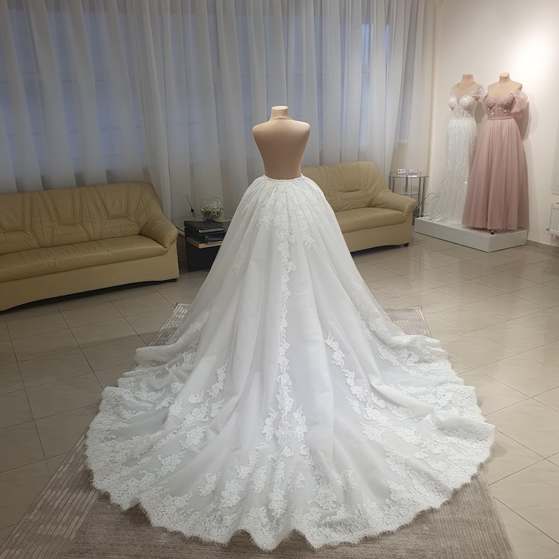 May include: A white wedding dress on a mannequin. The dress features a full skirt with intricate lace detailing along the hem and train. The dress is displayed in a bridal shop, with other gowns visible in the background.