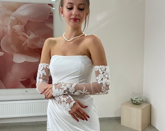 Puede incluir: Un vestido de novia blanco sin tirantes con una falda drapeada y una abertura alta. El vestido presenta mangas largas y transparentes adornadas con aplicaciones de encaje floral. Un collar de perlas completa el look. El vestido es elegante.