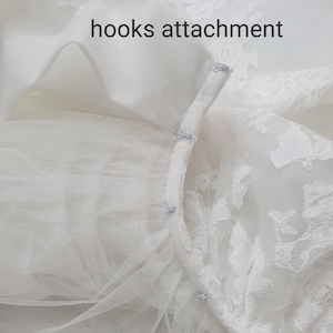 May include: Close-up of a white bridal gown detail. The image shows the hooks attachment of the dress, with delicate lace and tulle. The text "hooks attachment" is visible.