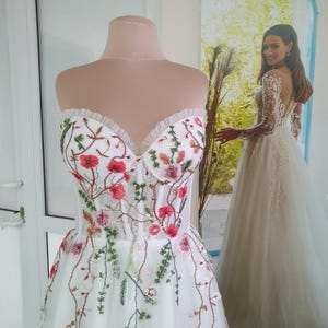 May include: A white strapless wedding dress with a tulle skirt and floral embroidery. The embroidery features pink, red, and green flowers.