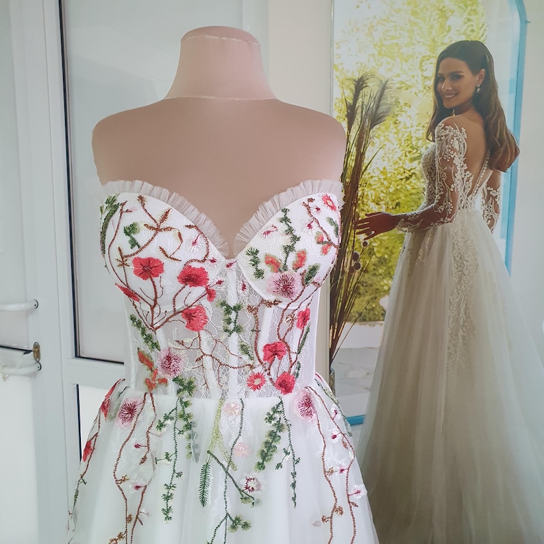 May include: A white strapless wedding dress with a tulle skirt and floral embroidery. The embroidery features pink, red, and green flowers.
